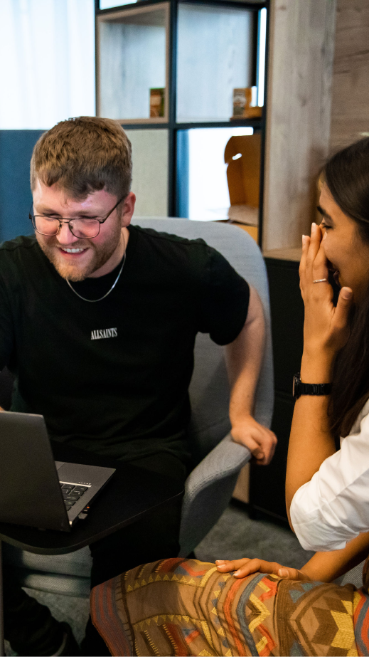 A woman and a man sitting and laughing with a laptop in front of them in an office.