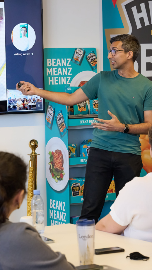 A man delivering a presentation to a group of people sitting down at tables.